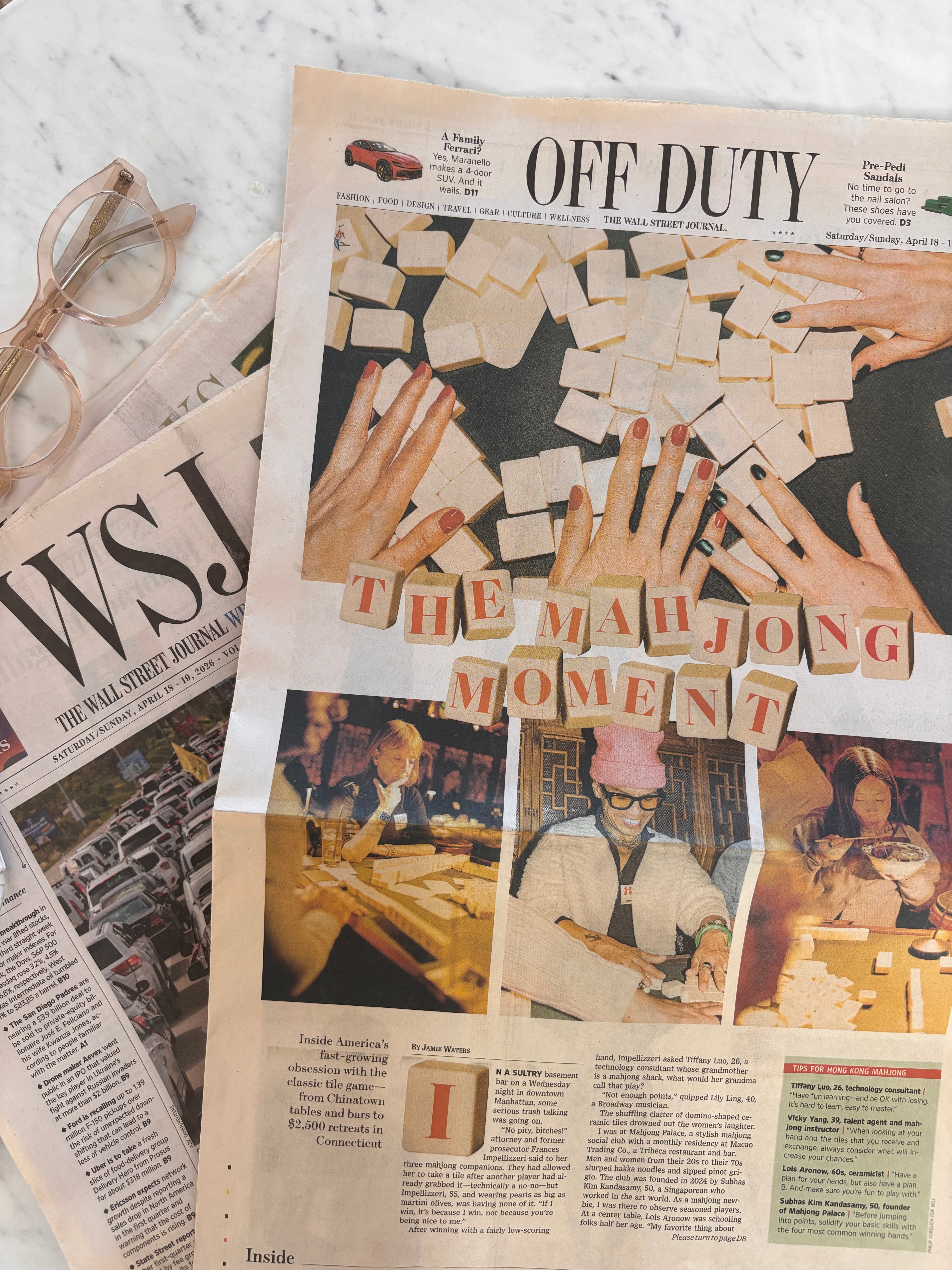 A styled flat-lay featuring a magazine or newspaper page with a large article titled "OFF DUTY" and a prominent cover photo about mahjong, alongside another folded newspaper page and a pair of eyeglasses on a light marble surface.